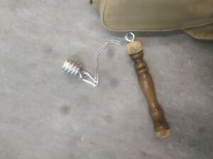 Un fléau dont le poids est en forme de cuillère à miel argenté, en aluminium. Le fléau est posé sur un sol gris, un sac apparaît en haut de l'image à ses côté