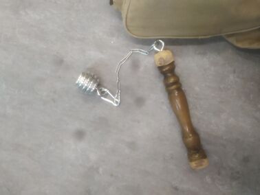 Un fléau dont le poids est en forme de cuillère à miel argenté, en aluminium. Le fléau est posé sur un sol gris, un sac apparaît en haut de l'image à ses côté